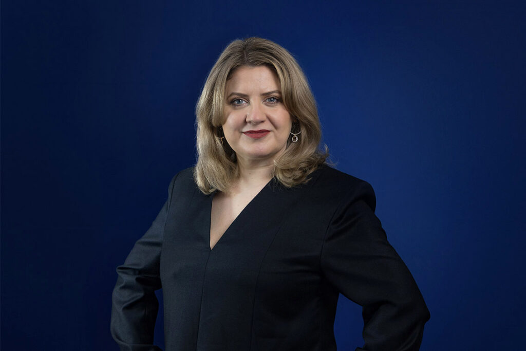 Porträt einer Frau mit blonden Haaren, die einen schwarzen Blazer trägt, vor einem blauen Hintergrund.